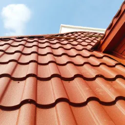 Czerwona dachówka modułowa na skośnym dachu w słoneczny dzień. Widoczne detale łączeń i faktura blachy, fragment budynku w tle i błękitne niebo z chmurą.