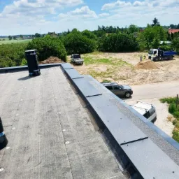 Wykończony dach pokryty papą z obróbkami blacharskimi w kolorze grafitowym. Widok z góry na tle zieleni, nieba i budowy.