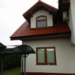 Czerwona dachówka na białym domu z brązowymi oknami. Widoczny fragment zadaszenia nad wejściem i klimatyzator na ścianie. Pochmurny dzień.