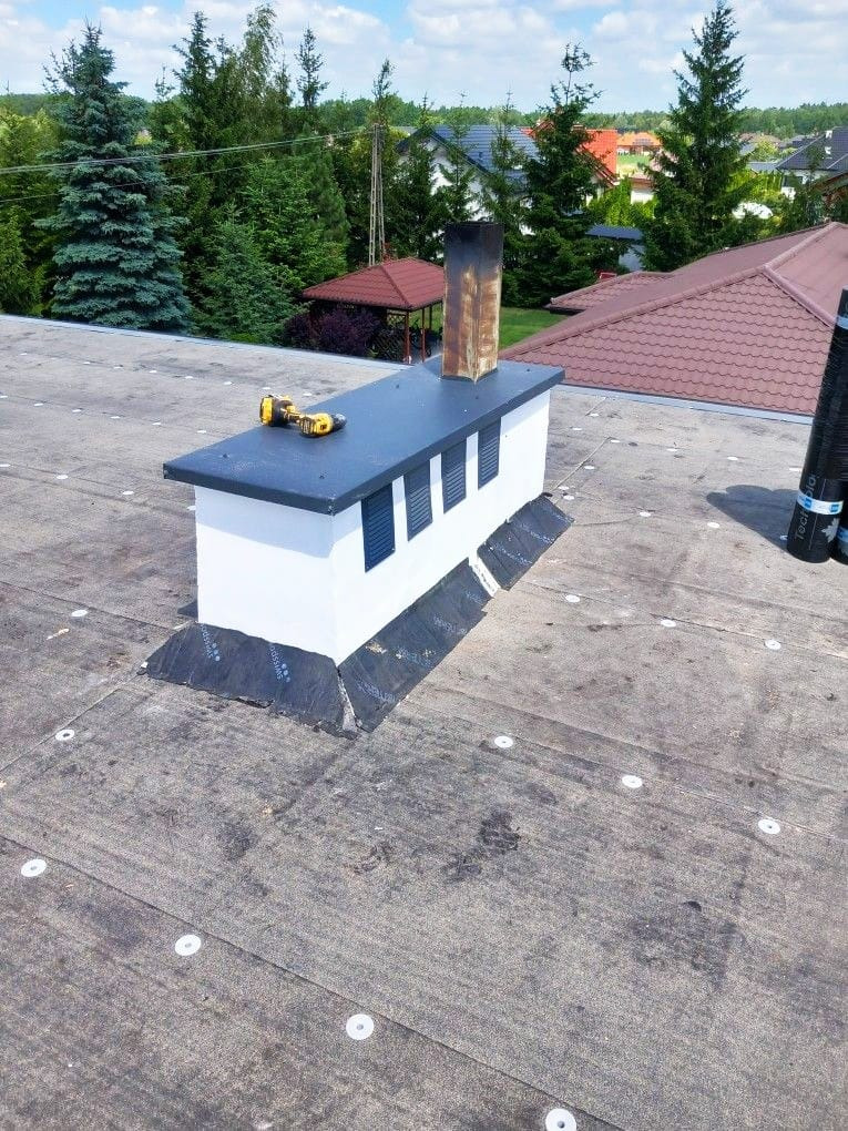 Wykończony komin wentylacyjny na dachu pokrytym papą. Widoczne narzędzia i obróbki blacharskie. W tle drzewa i zabudowania.