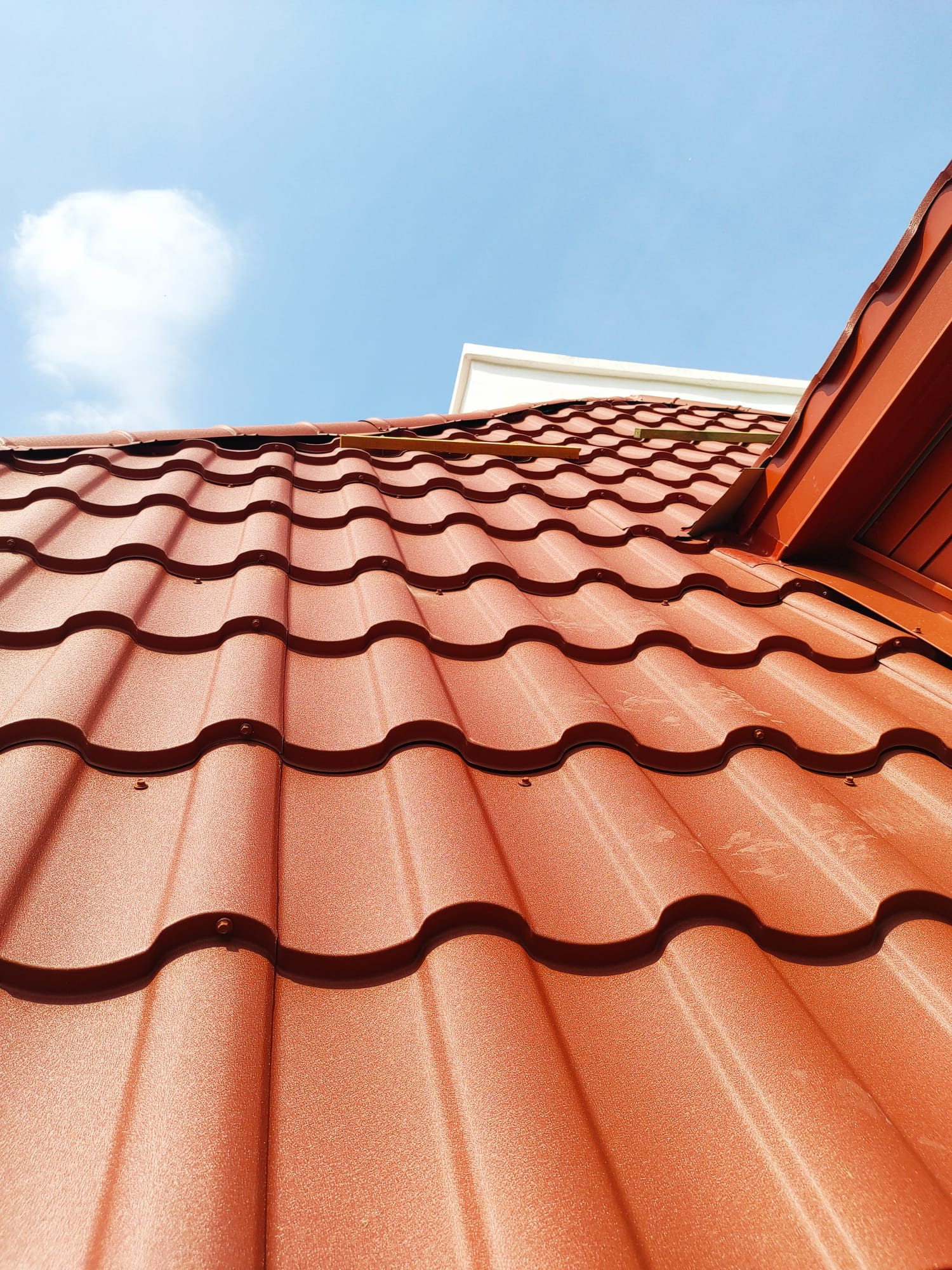 Czerwona dachówka modułowa na skośnym dachu w słoneczny dzień. Widoczne detale łączeń i faktura blachy, fragment budynku w tle i błękitne niebo z chmurą.