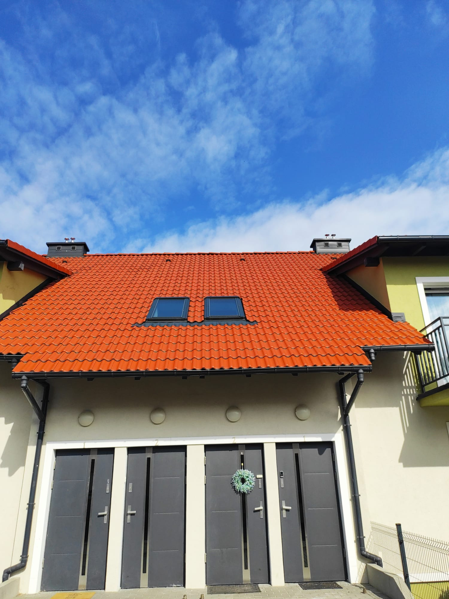 Czerwony dach z dwoma oknami dachowymi na tle błękitnego nieba. Trzy szare drzwi wejściowe z wieńcem na jednych. Nowoczesna architektura.