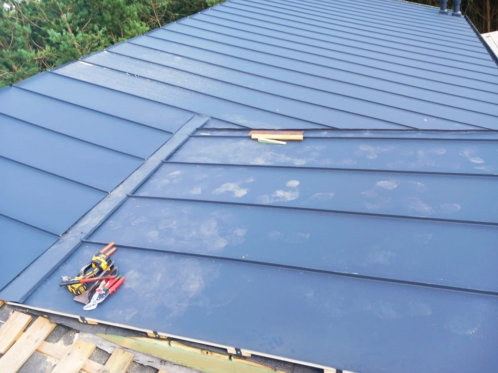 Niebieski dach z blachy na rąbek stojący w trakcie montażu, widoczne narzędzia dekarskie i fragment więźby. Ujęcie z góry, z widocznym fragmentem lasu w tle.