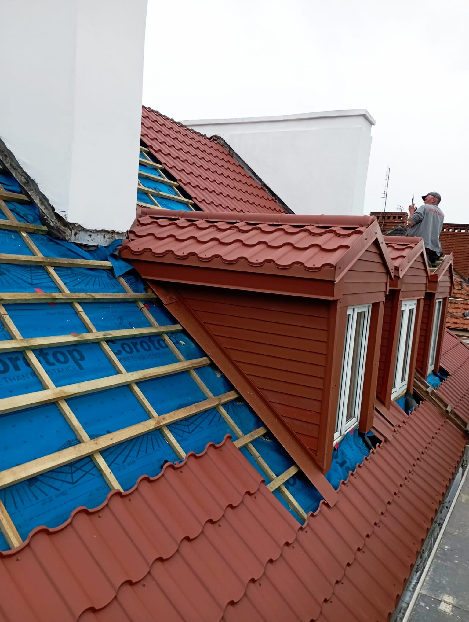 Montaż brązowej blachodachówki na dachu domu z oknami połaciowymi. Widoczna membrana dachowa i drewniana konstrukcja. Prace dekarskie w toku.