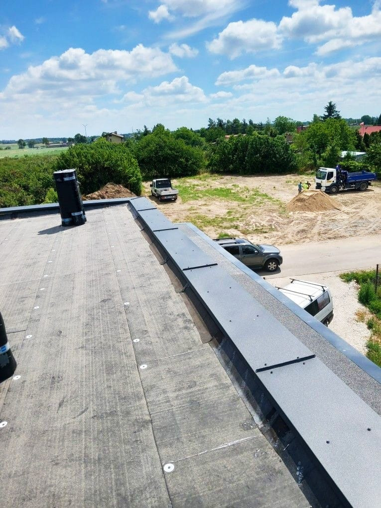 Wykończony dach pokryty papą z obróbkami blacharskimi w kolorze grafitowym. Widok z góry na tle zieleni, nieba i budowy.
