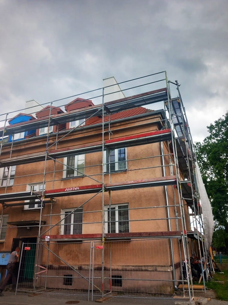 Budynek w rusztowaniach podczas prac remontowych. Widoczne elementy dachu i elewacji. Perspektywa z dołu, pochmurne niebo w tle. Prace w toku.