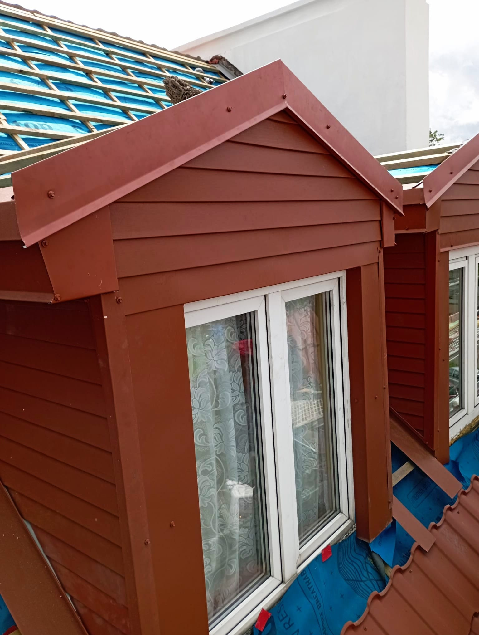 Brunatny lukarna z oknem na tle błękitnej membrany dachowej i fragmentu dachu z czerwonej dachówki. Widoczne elementy konstrukcji i obróbki blacharskiej.