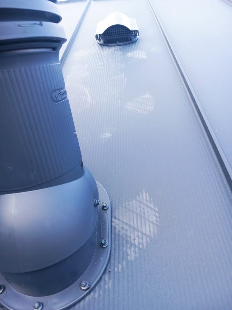 Szary, blaszany dach z kominkiem wentylacyjnym i wywietrznikiem. Widok z dołu, ukazujący detale montażu i fakturę blachy.