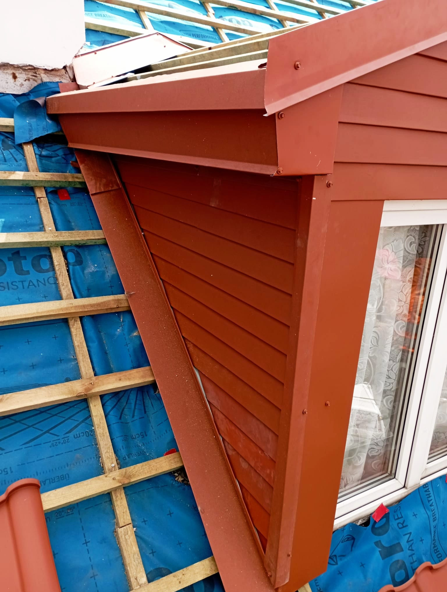 Czerwona obróbka blacharska lukarny z oknem, widoczna membrana dachowa i drewniana konstrukcja dachu. Detal wykończenia dachu w bliskim ujęciu.