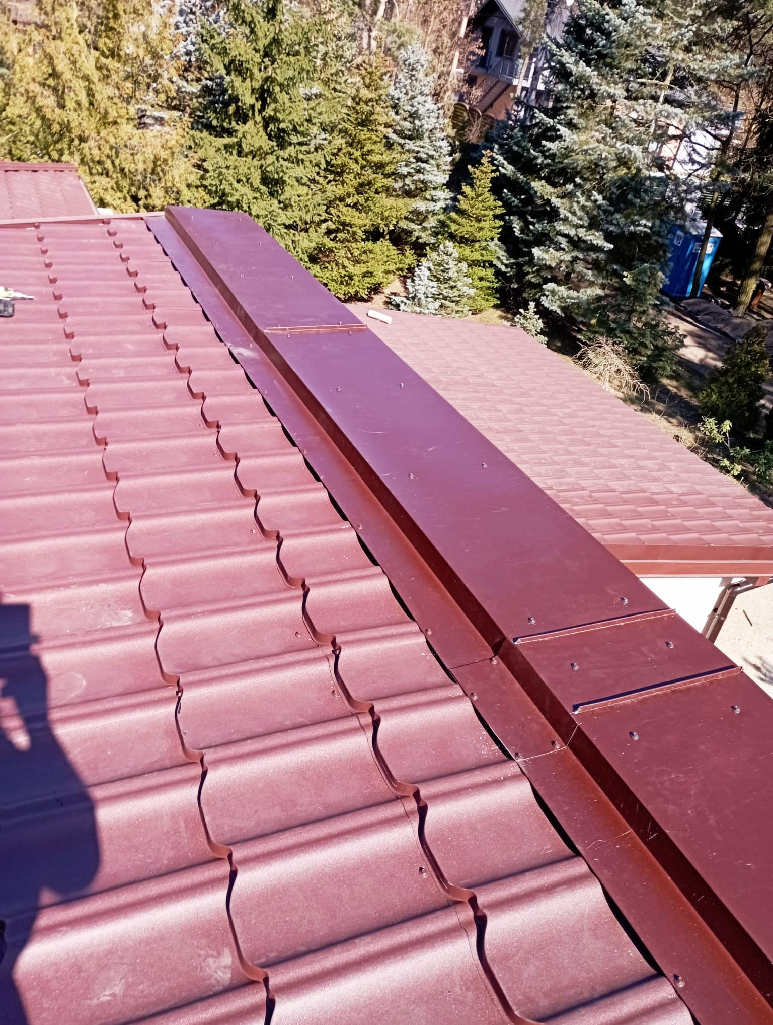 Czerwono-brązowy dach z blachodachówki modułowej, z widoczną obróbką blacharską w kolorze dachu. W tle drzewa iglaste i budynek.
