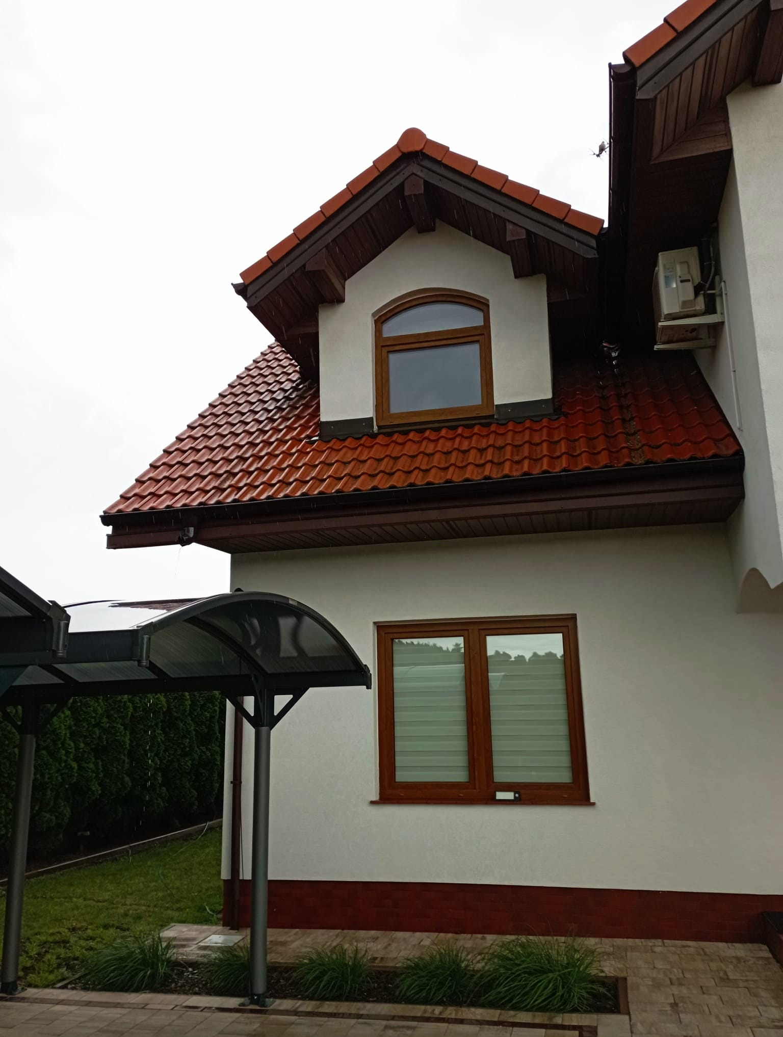 Czerwona dachówka na białym domu z brązowymi oknami. Widoczny fragment zadaszenia nad wejściem i klimatyzator na ścianie. Pochmurny dzień.