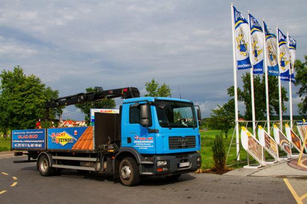 Błękitna ciężarówka z dźwigiem, załadowana dachówką, zaparkowana obok rzędu flag reklamowych z logo firmy, na tle pochmurnego nieba.