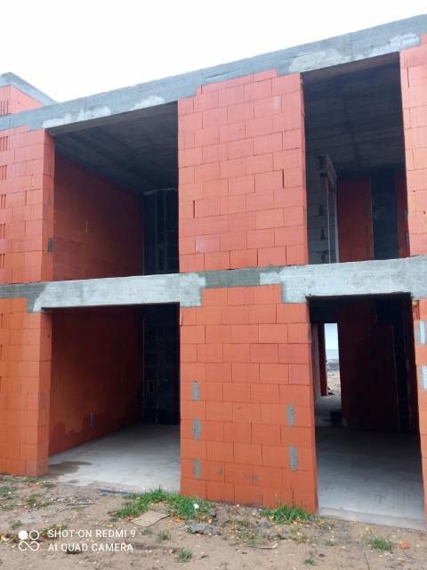 Dwukondygnacyjny budynek w trakcie budowy z czerwonej cegły, widoczne surowe otwory okienne i drzwiowe, szary betonowy wieniec, teren wokół budynku częściowo porośnięty trawą.