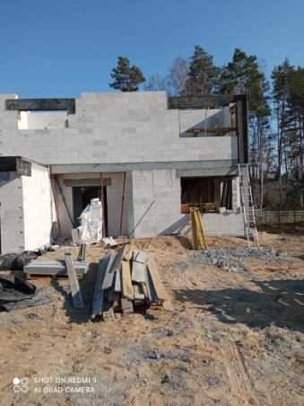 Budowa domu z betonu komórkowego w stanie surowym otwartym, widoczna sterta materiałów budowlanych i drabina oparta o ścianę, las w tle.