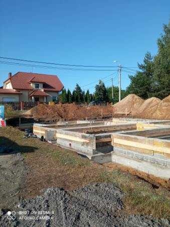 Fundamenty pod budowę domu jednorodzinnego, widoczne szalunki i zbrojenie, w tle hałda ziemi i istniejący dom z czerwoną dachówką.