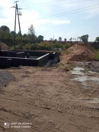 Wylany fundament pod budynek jednorodzinny z widocznym zbrojeniem, na działce z hałdą piasku i słupem wysokiego napięcia w tle.