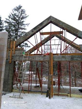 Surowa konstrukcja domu w trakcie budowy z widocznym szkieletem dachu, metalowymi wspornikami i drabiną na tle zimowego krajobrazu.