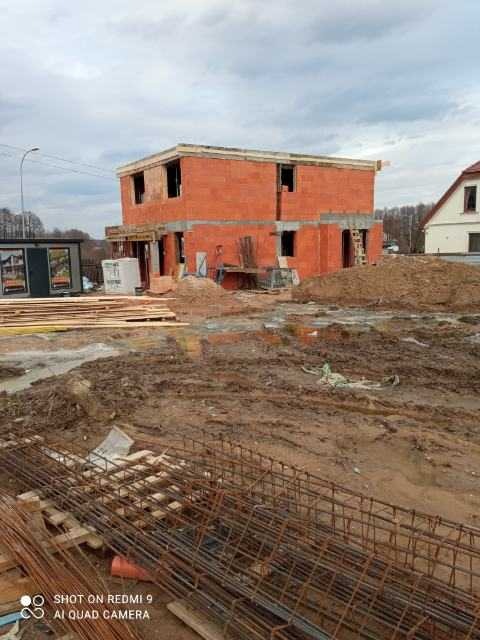 Surowy stan budowy dwukondygnacyjnego domu z czerwonej cegły, widoczne zbrojenia i materiały budowlane na błotnistym terenie w pochmurny dzień.