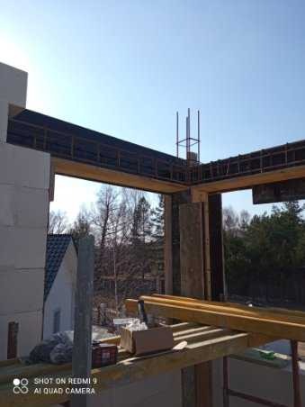 Wznoszenie surowej konstrukcji budynku z betonu komórkowego, widoczne drewniane szalunki i stalowe zbrojenia, w tle zieleń drzew.