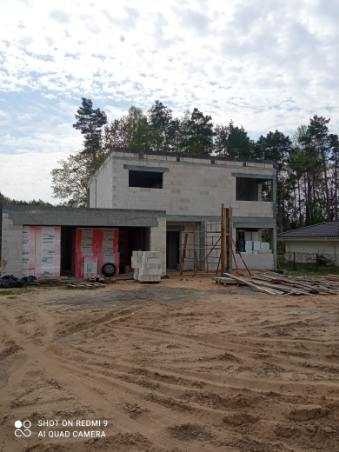 Surowy stan dwukondygnacyjnego budynku z bloczków betonowych w trakcie budowy, widoczne rusztowania i materiały budowlane na placu budowy.