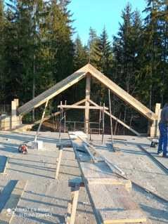 Konstrukcja drewniana dachu w trakcie budowy domu murowanego, widoczne elementy więźby, rusztowanie, betonowa płyta fundamentowa i las w tle.