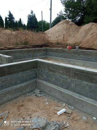 Wykopany fundament z bloczków betonowych, częściowo zalany betonem, z widoczną stertą piasku w tle.