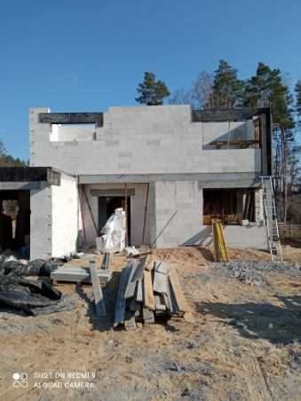 Budowa domu z betonu komórkowego: surowy stan, widoczne bloczki, rusztowanie i materiały budowlane na placu budowy, drzewa w tle.