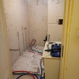 Oleksandr Kachkar GREENINDI - Łazienka w trakcie demontażu płytek, z odsłoniętymi rurami, narzędziami glazurniczymi na prowizorycznym blacie i widocznymi śladami po skuciu starej okładziny na ścianach.