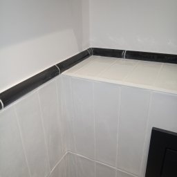 Oleksandr Kachkar GREENINDI - Białe płytki ścienne z czarnym, błyszczącym dekorem i czarnym przyciskiem spłuczki w narożniku łazienki. Widoczny fragment instalacji wodnej.