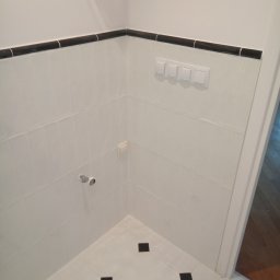 Oleksandr Kachkar GREENINDI - Białe płytki z czarnym pasem dekoracyjnym i czarnymi akcentami na podłodze w rogu łazienki. Widoczne włączniki i instalacja wodna.