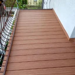 Balkon z brązową deską kompozytową, widok z góry. Metalowa balustrada z dekoracyjnymi elementami. Tło: budynek i zieleń. Układ poziomy desek.
