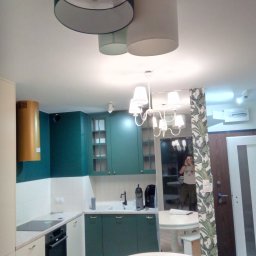 Oleksandr Kachkar GREENINDI - Nowoczesna kuchnia z zielonymi szafkami, białymi blatami i okrągłymi stołami, oświetlona wiszącymi lampami z abażurami, widok z perspektywy osoby robiącej zdjęcie w lustrze.