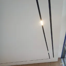 Biały sufit z dwoma czarnymi, podłużnymi lampami wpuszczonymi, z widocznymi panelami drewnianymi na dole kadru.