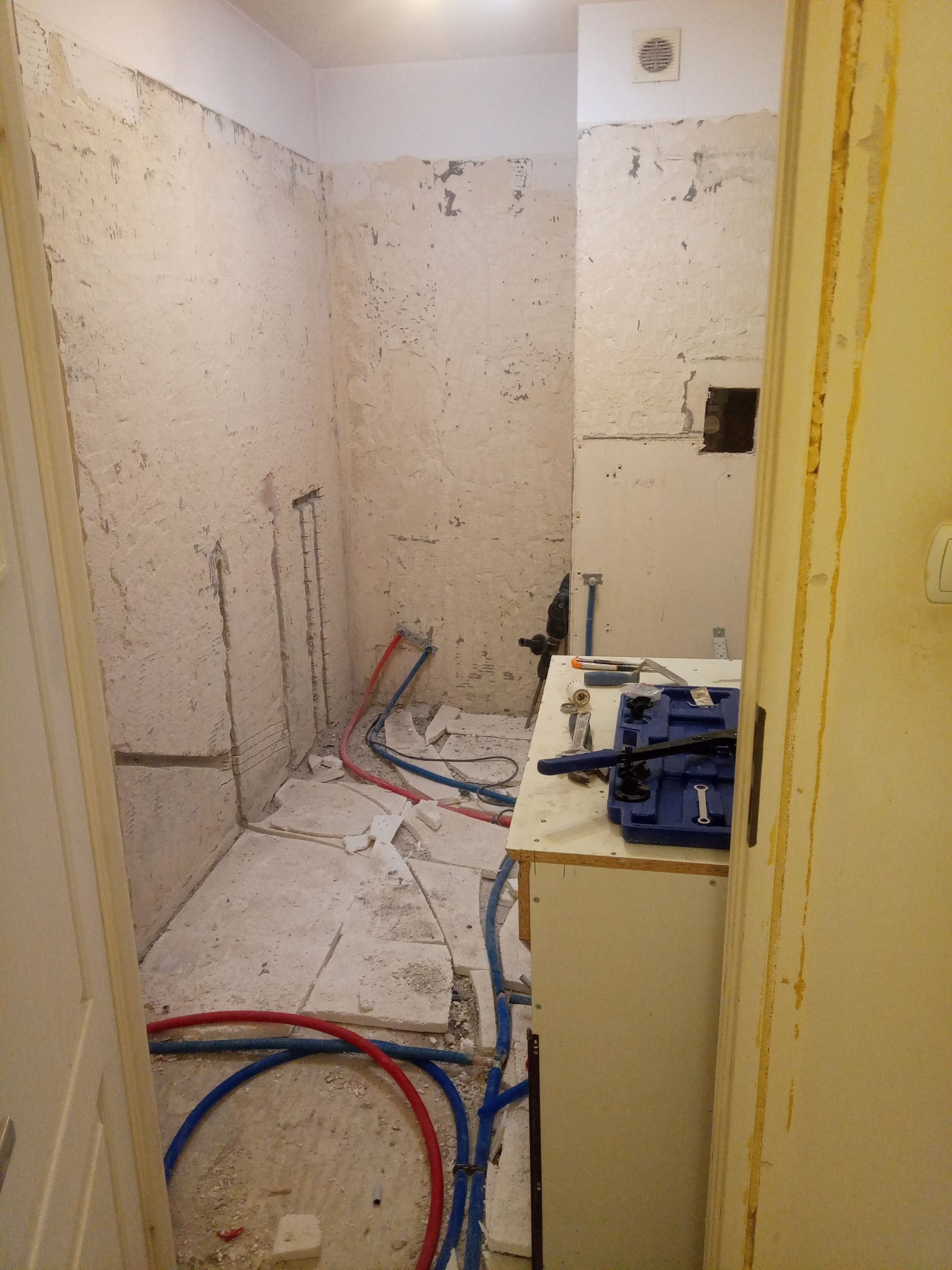 Łazienka w trakcie demontażu płytek, z odsłoniętymi rurami, narzędziami glazurniczymi na prowizorycznym blacie i widocznymi śladami po skuciu starej okładziny na ścianach.