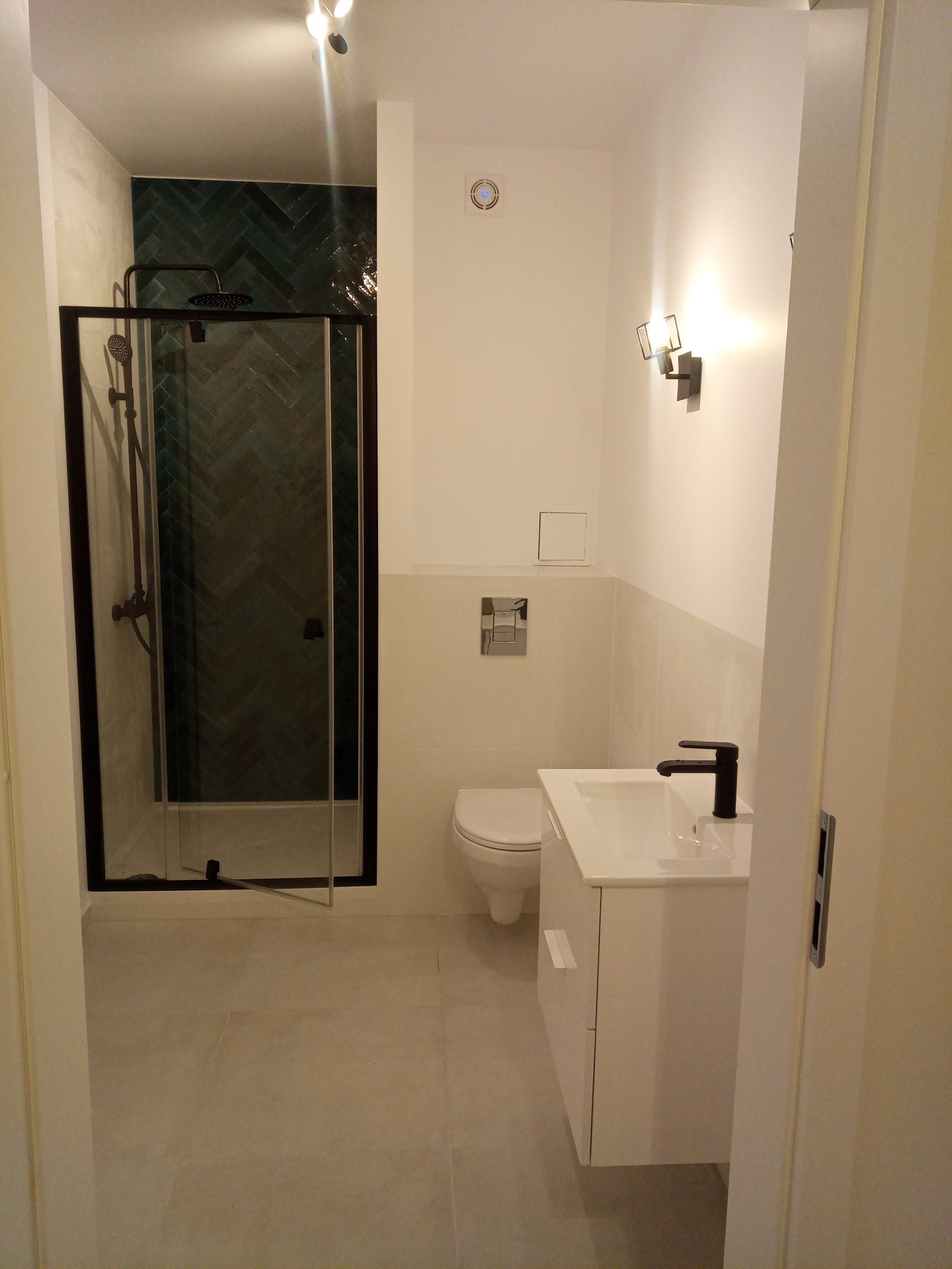 Nowoczesna łazienka z kabiną prysznicową z czarną ramą i armaturą, zielonymi płytkami ułożonymi w jodełkę, białą umywalką z szafką i czarną baterią.