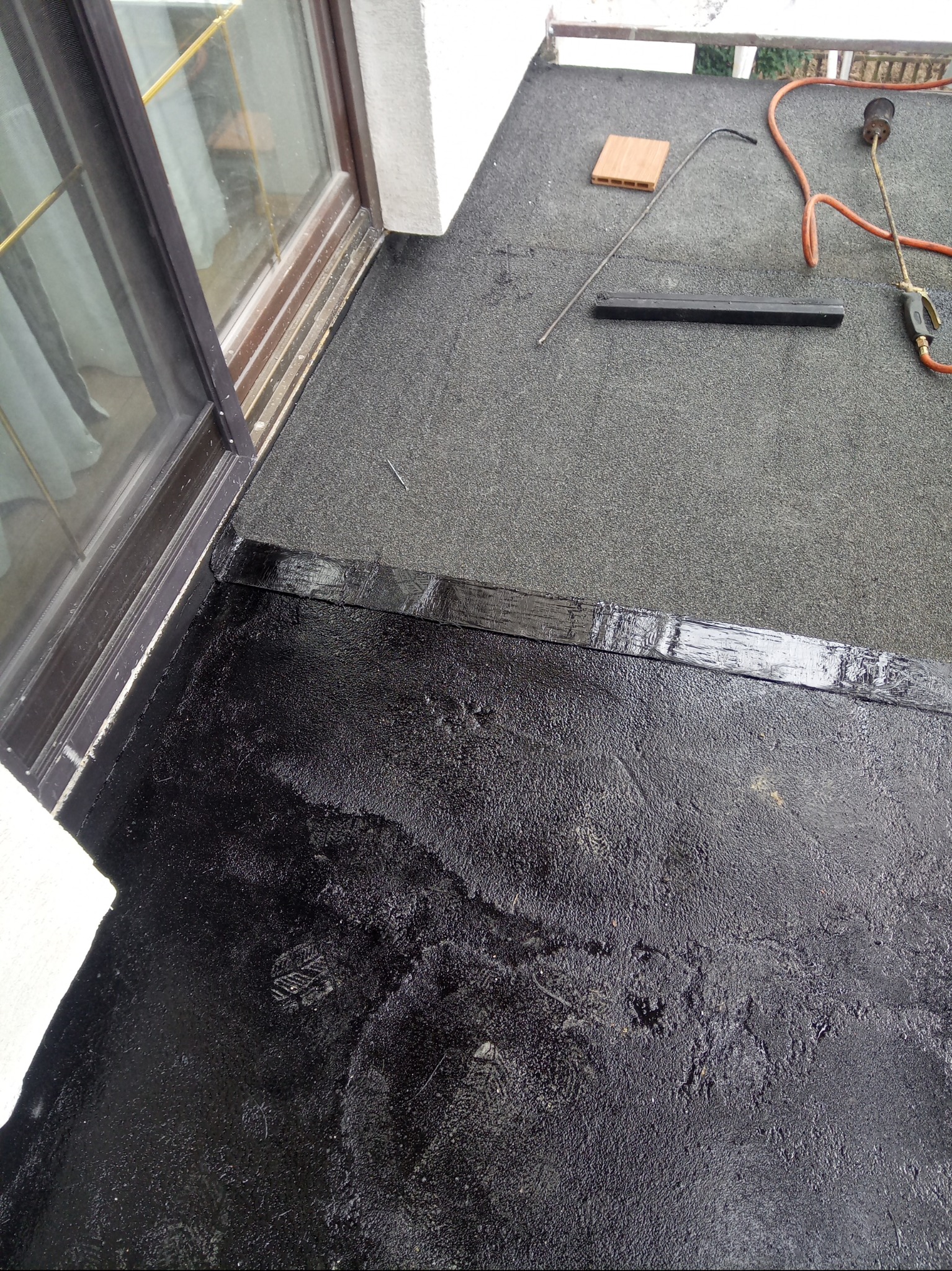 Czarną hydroizolacja na balkonie z widocznymi narzędziami: palnik, listwa i cegła. Częściowo pokryta, mokra powierzchnia. Detal prac wykończeniowych na zewnątrz.