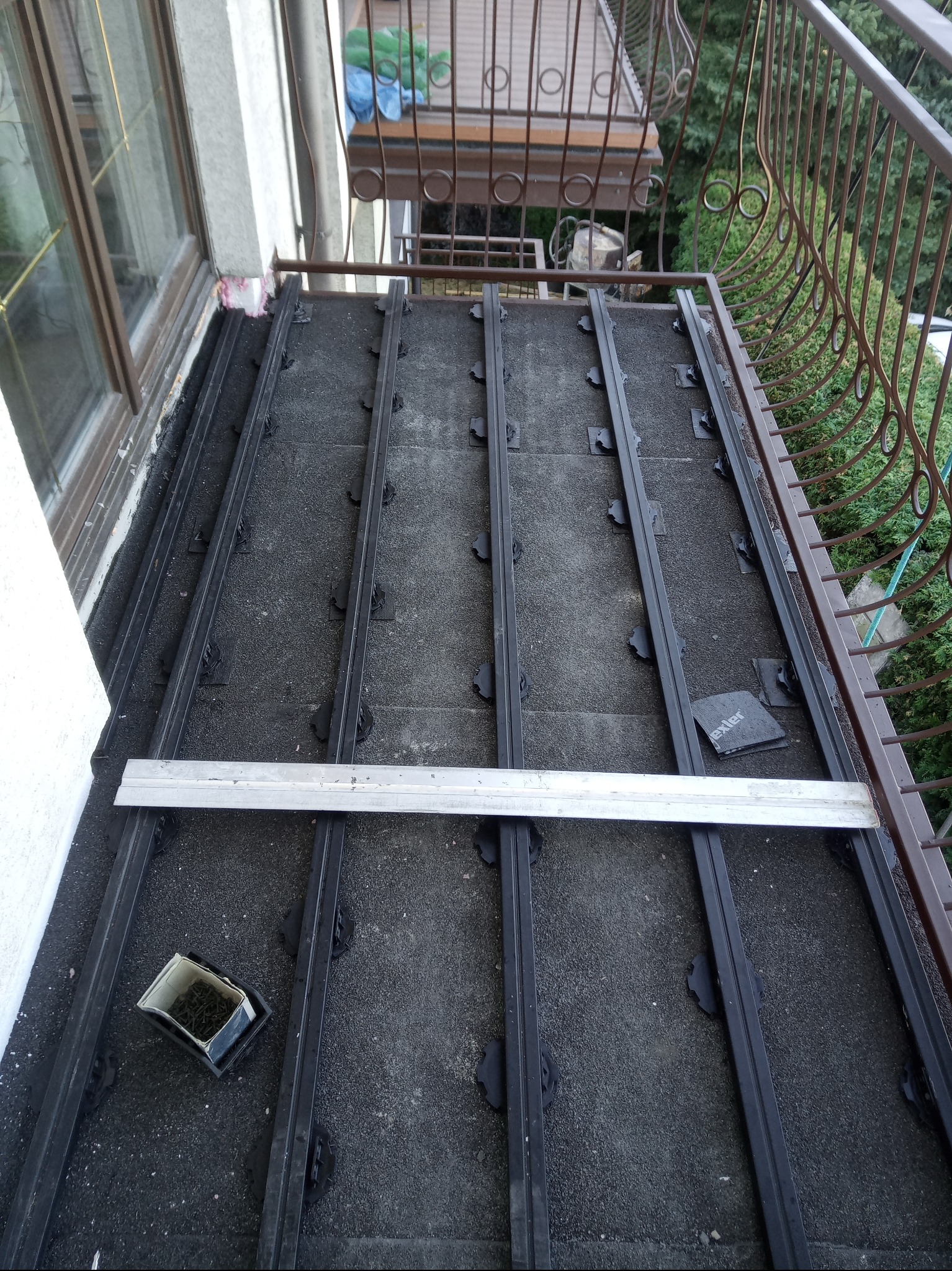 Montaż legarów na balkonie z widocznymi elementami mocującymi i aluminiową łatą. Przygotowanie do ułożenia desek podłogowych na tarasie.