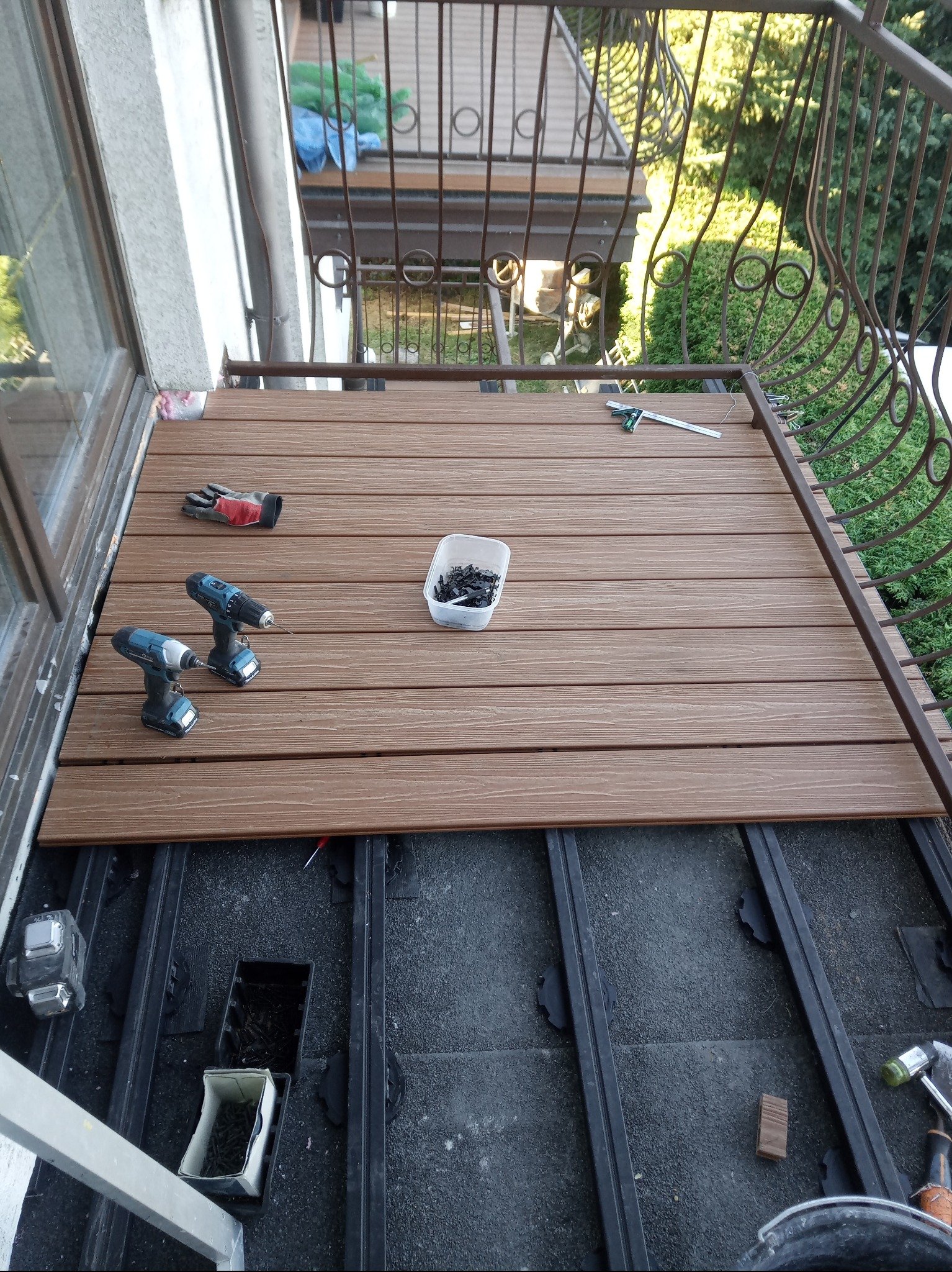 Montaż deski kompozytowej na balkonie. Widoczne narzędzia, wkrętarka, poziomica i rękawice robocze. Część desek już ułożona, pozostałe czekają na montaż.