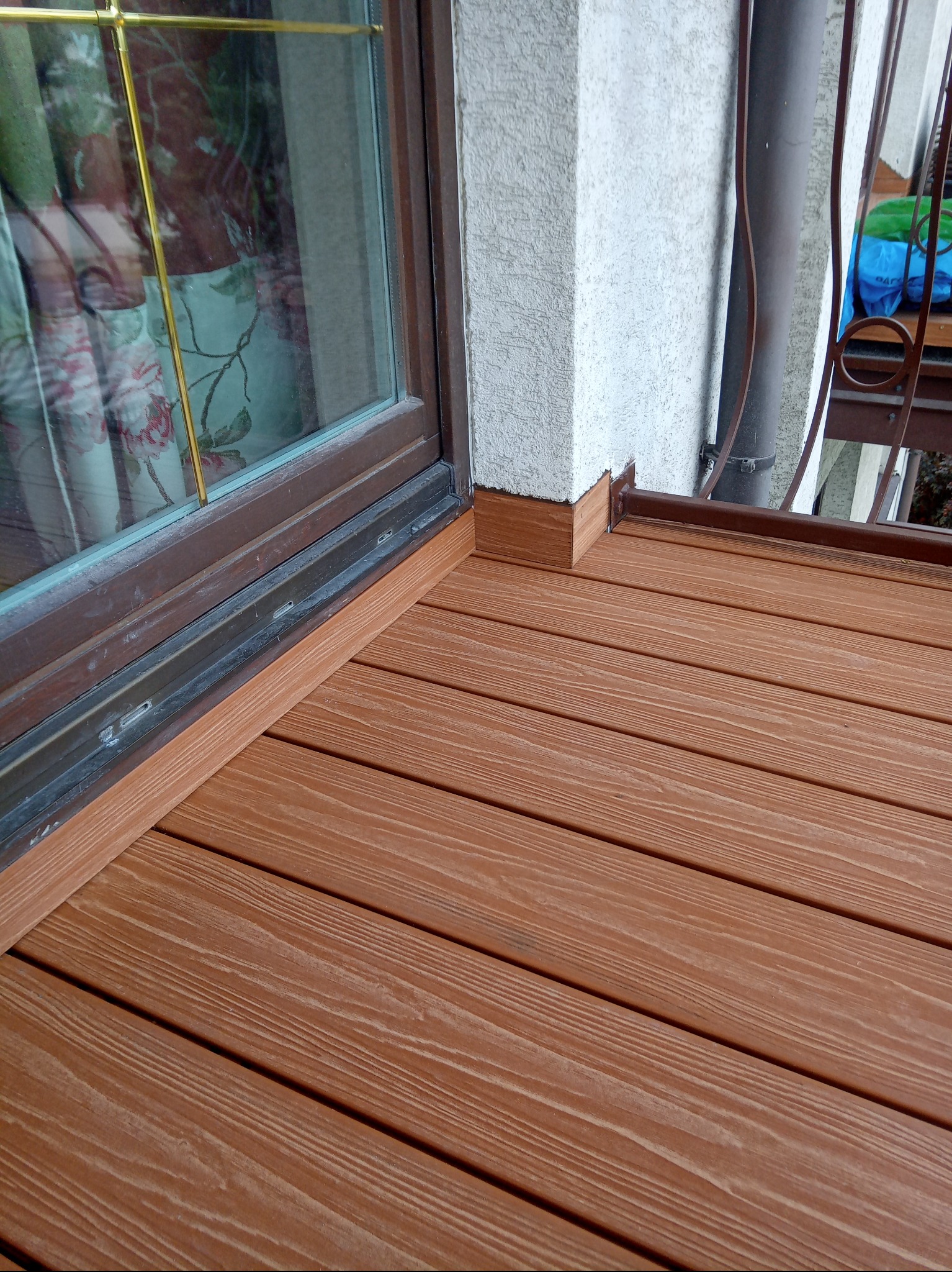Wykończony taras z desek kompozytowych w odcieniach brązu, widoczny przy oknie i narożniku budynku. Detal łączenia desek z elewacją i ramą okienną.