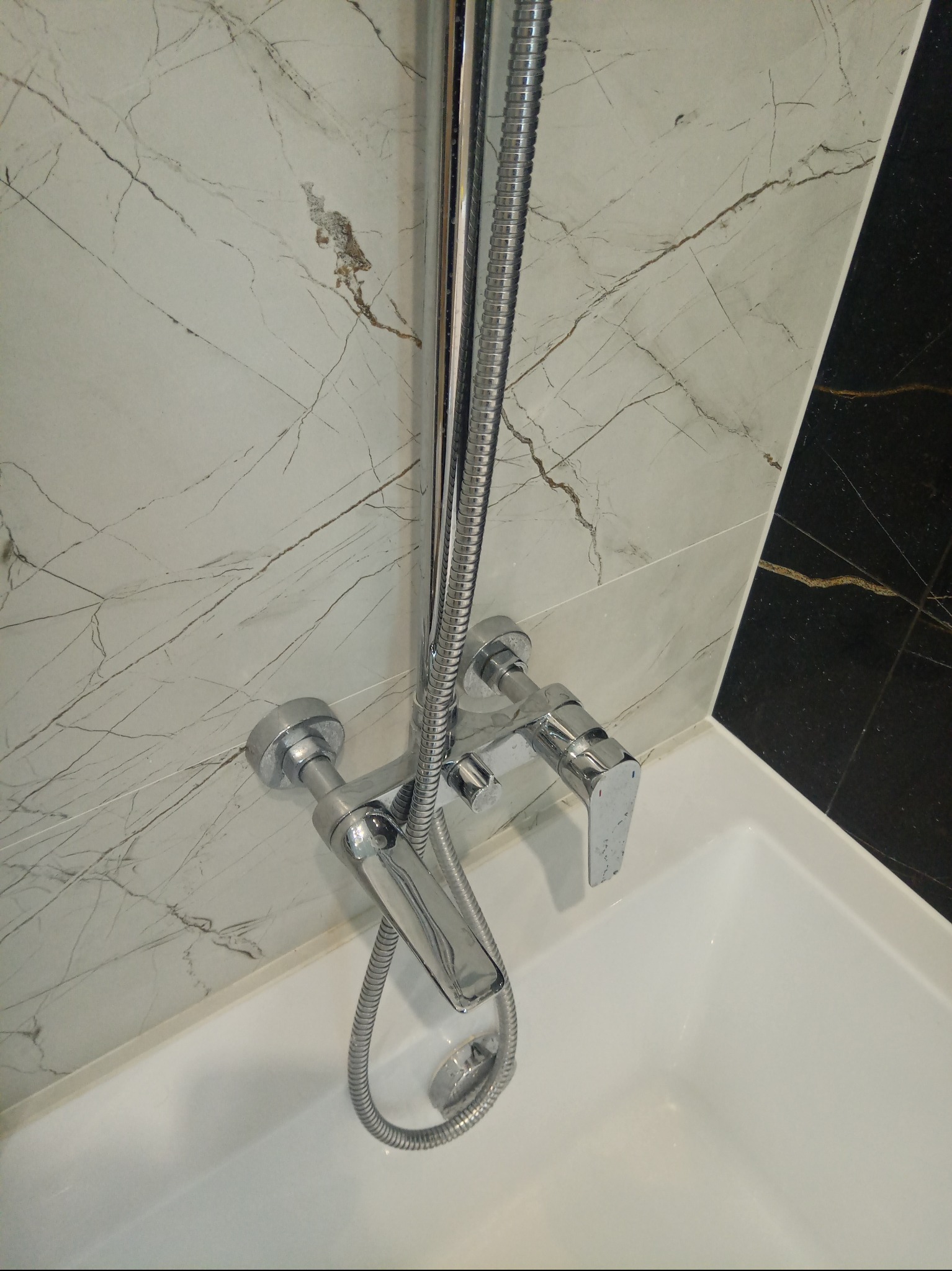 Bateria wannowa chromowana z deszczownicą na tle marmurowych płytek w odcieniach bieli i czerni. Nowoczesna armatura łazienkowa po montażu.