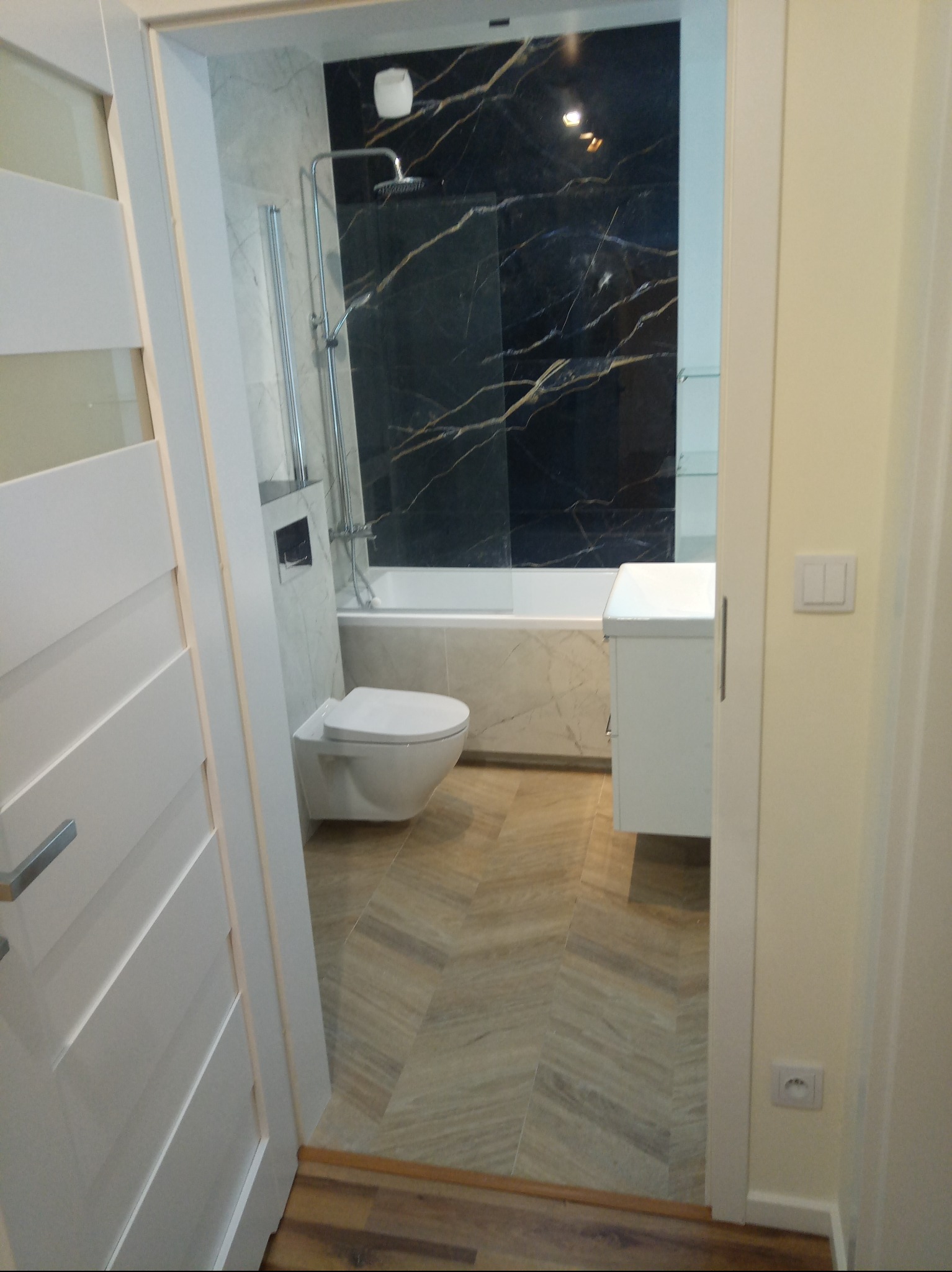 Nowoczesna łazienka z czarną ścianą imitującą marmur, białą armaturą i podłogą ułożoną w jodełkę, widoczna zza uchylonych białych drzwi.