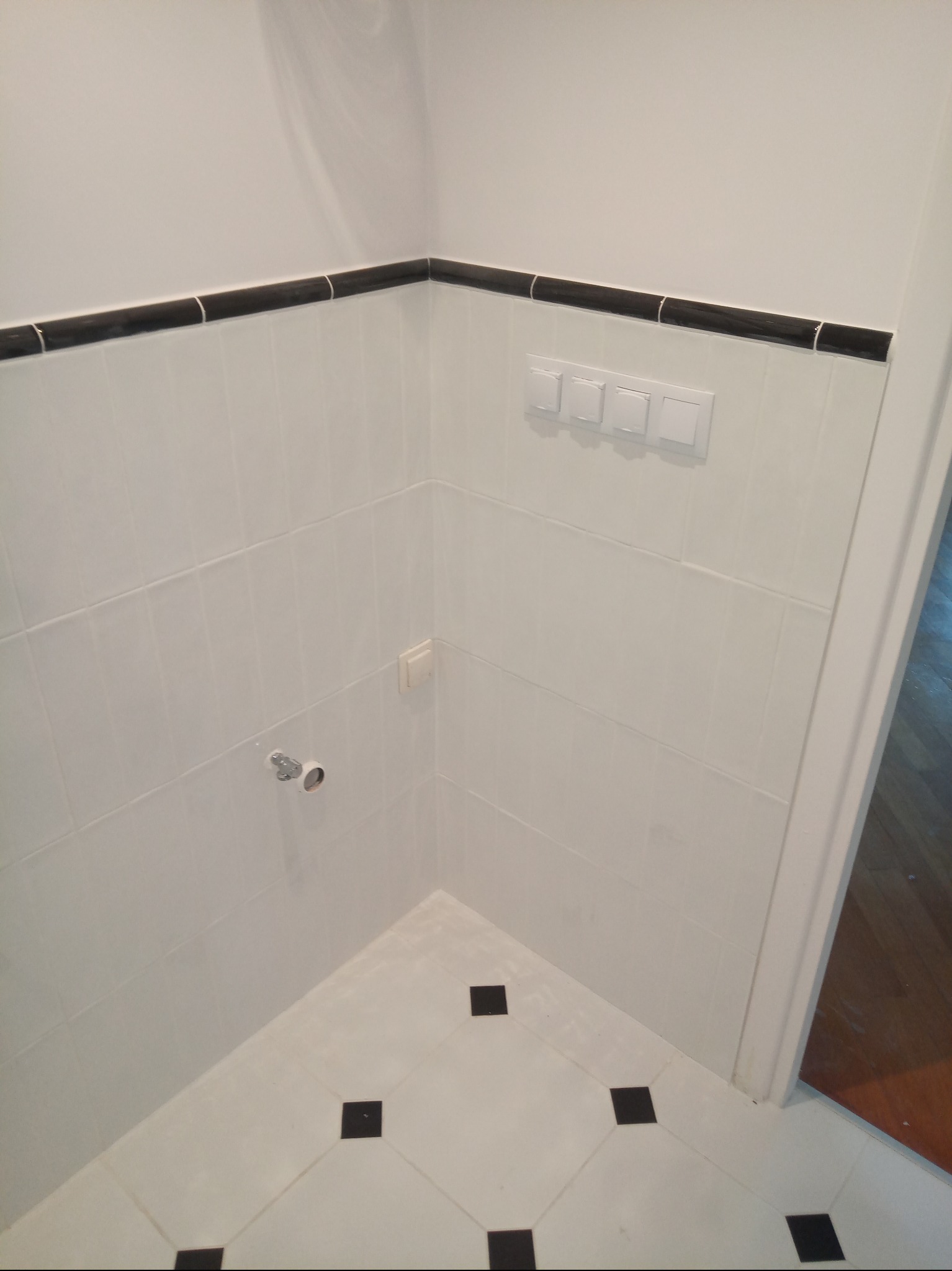 Białe płytki z czarnym pasem dekoracyjnym i czarnymi akcentami na podłodze w rogu łazienki. Widoczne włączniki i instalacja wodna.
