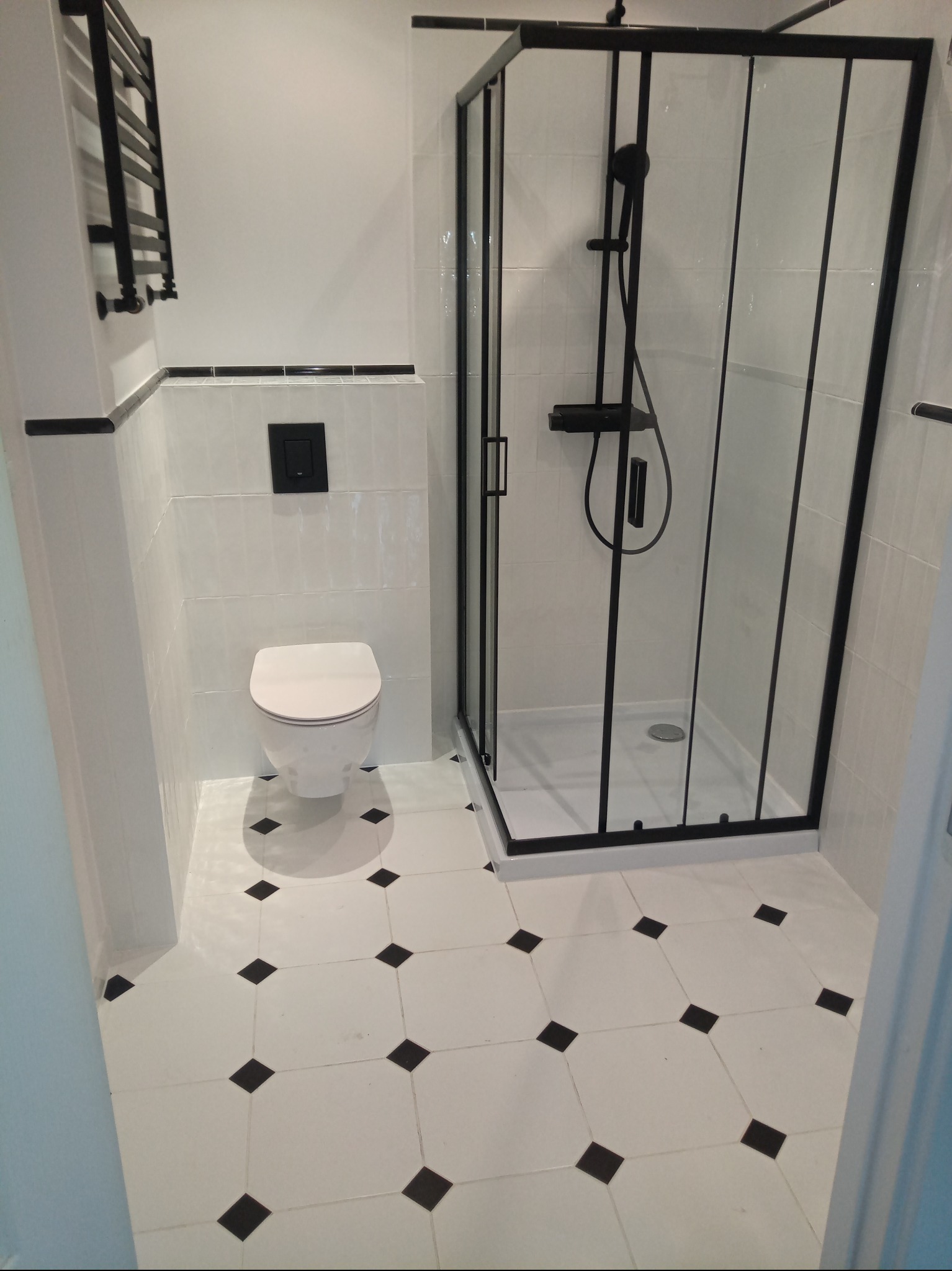 Biała łazienka z czarnymi akcentami: kabina prysznicowa, grzejnik, toaleta. Podłoga w biało-czarną szachownicę. Minimalistyczny design, nowoczesne wykończenie.