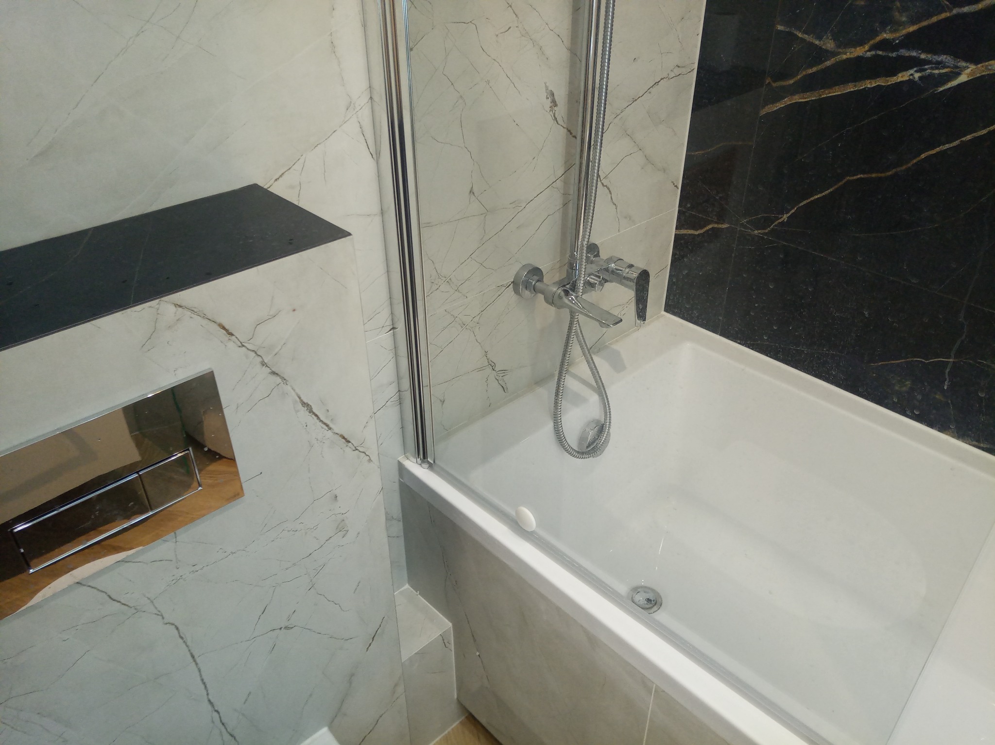 Wykończona łazienka z wanną, szklanym parawanem i baterią. Ściany wyłożone marmurowymi płytkami w odcieniach bieli i czerni. Widoczny panel sterujący spłuczką.