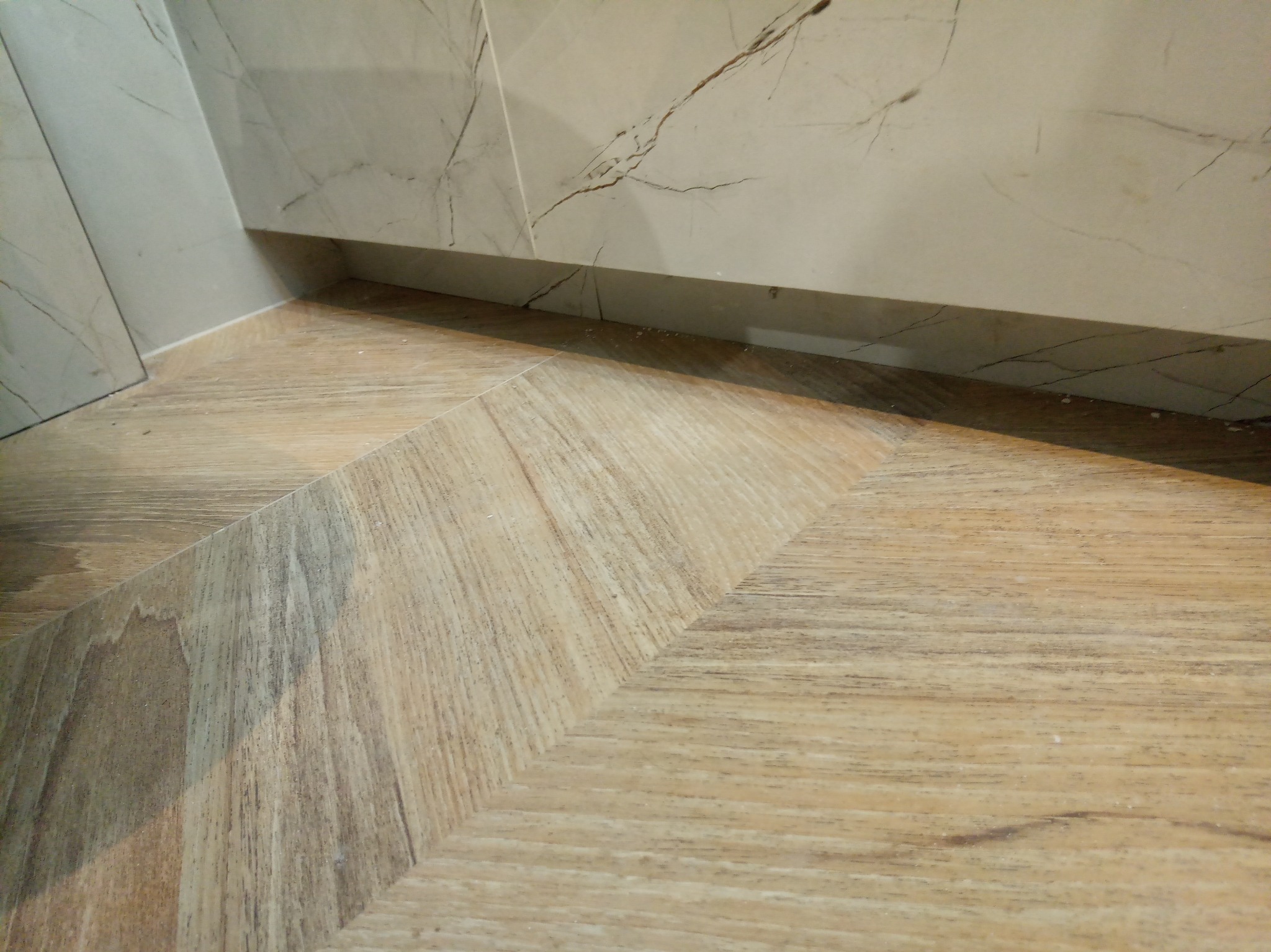 Ukończona podłoga z paneli ułożonych we wzór jodełki, z widoczną ścianą wykończoną płytkami imitującymi marmur, prezentująca detale wykończenia wnętrza.