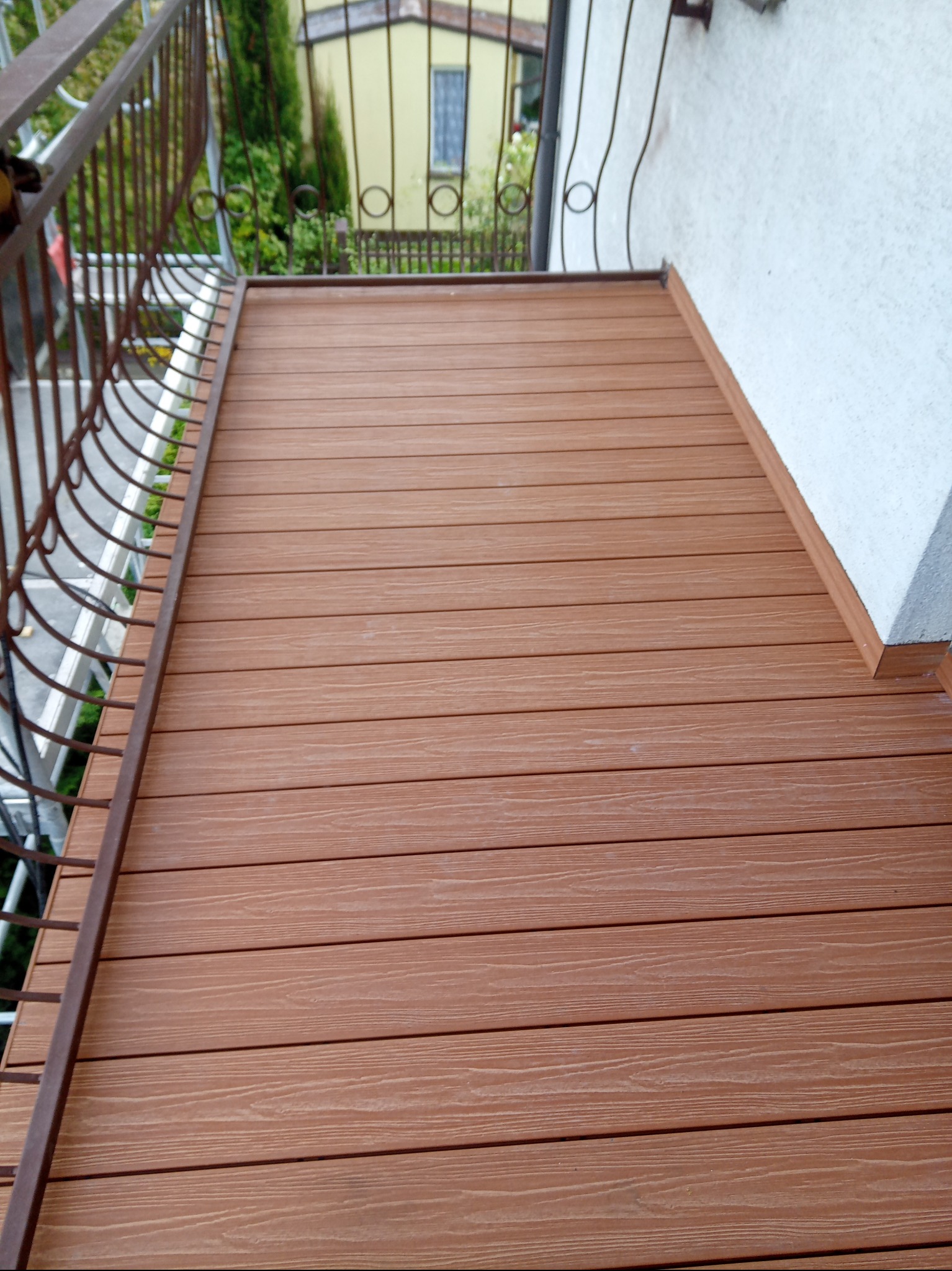 Balkon z brązową deską kompozytową, widok z góry. Metalowa balustrada z dekoracyjnymi elementami. Tło: budynek i zieleń. Układ poziomy desek.