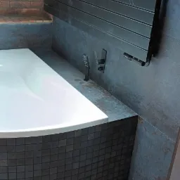 Nowoczesna łazienka na poddaszu z białą wanną obudowaną mozaiką, grafitowymi płytkami ściennymi, czarnym grzejnikiem drabinkowym i drewnianą podłogą, oświetlona ciepłym światłem spod wanny.