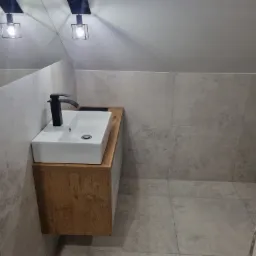 Nowoczesna łazienka na poddaszu z umywalką nablatową na drewnianej szafce, czarną armaturą i szarymi płytkami imitującymi kamień.