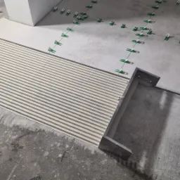 Przygotowanie podłoża pod płytki z nałożonym klejem w regularne paski i systemem poziomowania płytek z zielonymi klipsami, widoczny fragment ściany i drabina.