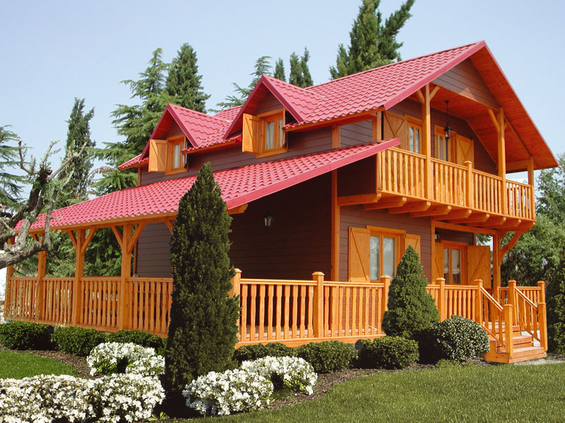 Dwukondygnacyjny dom z bali z czerwonym dachem, drewnianymi balkonami i werandą, otoczony zielonym trawnikiem i ozdobnymi krzewami.